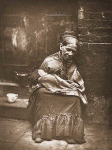 A lady sits on a step, a shawl wrapped around her shoulders, a bundle on her lap.