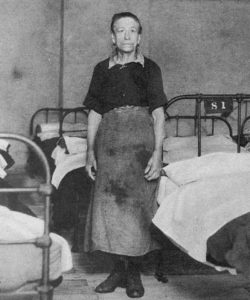 A female attendant stands by a bed at a common lodging house.